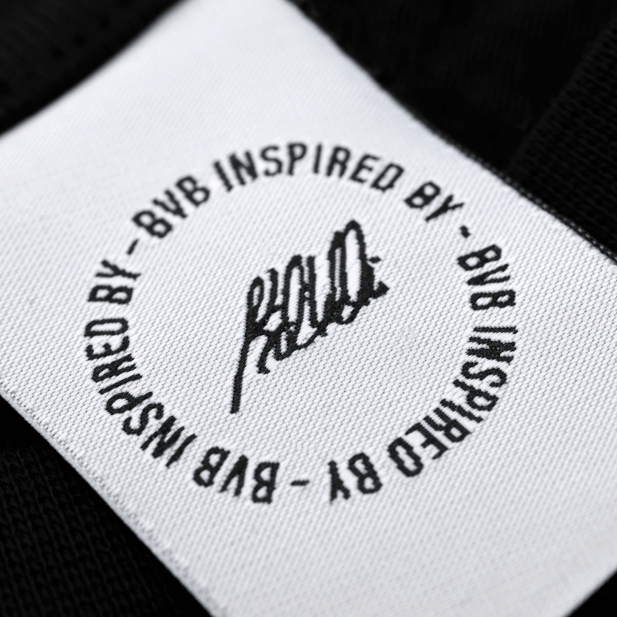 Camiseta negra del BVB INBY III con un estampado circular «BVB Inspired By» como detalle en una etiqueta de tela blanca, producto oficial del Borussia Dortmund.