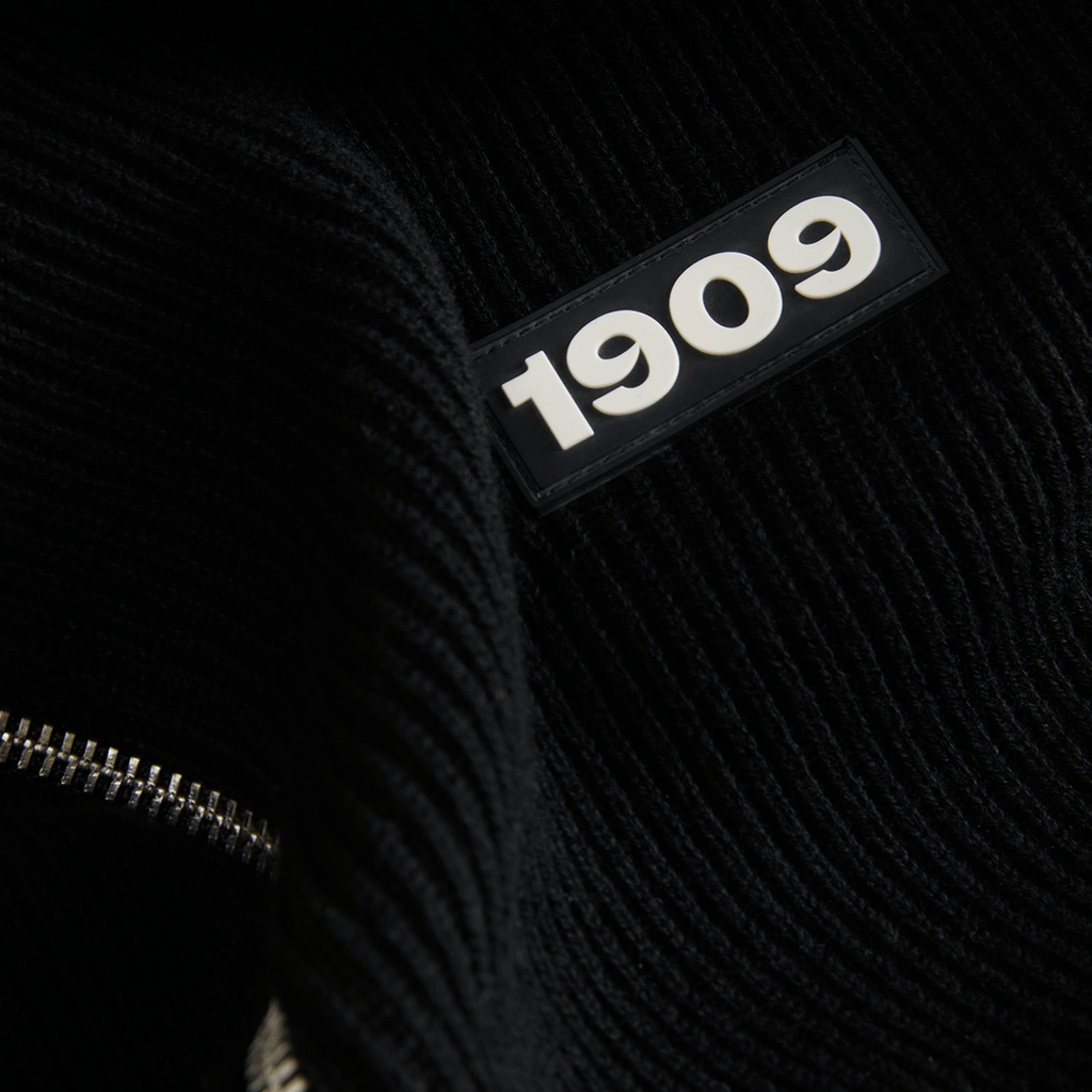 Schwarzer Strickstoff mit eingeprägtem 1909 Schriftzug und sichtbarem Reißverschluss in Nahaufnahme.