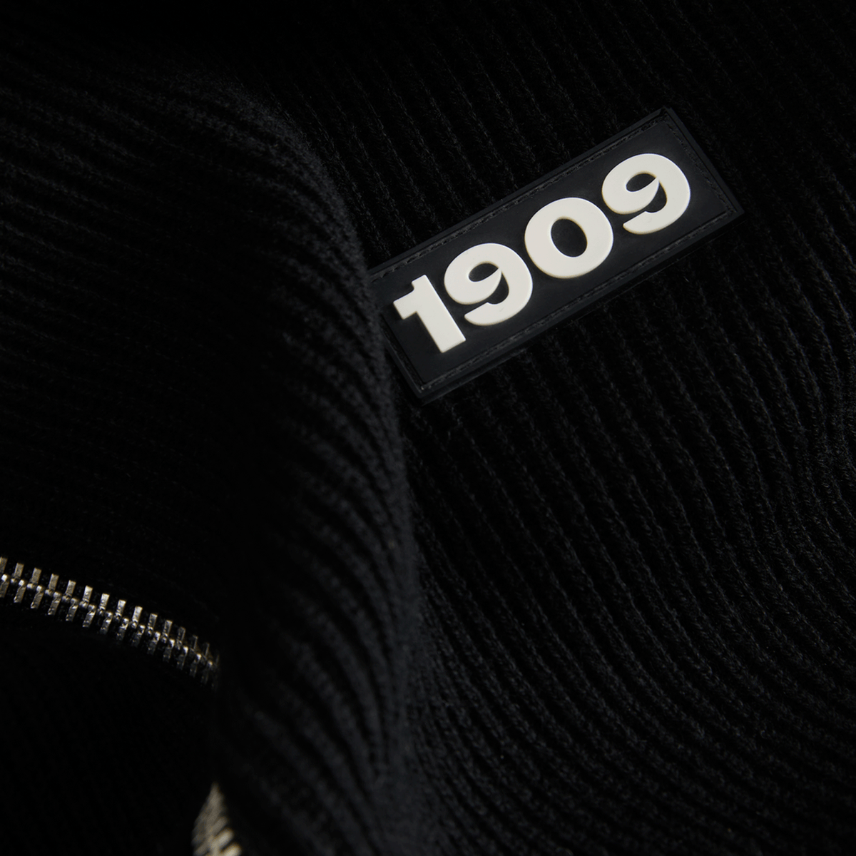 Schwarzer Strickstoff mit eingeprägtem 1909 Schriftzug und sichtbarem Reißverschluss in Nahaufnahme.