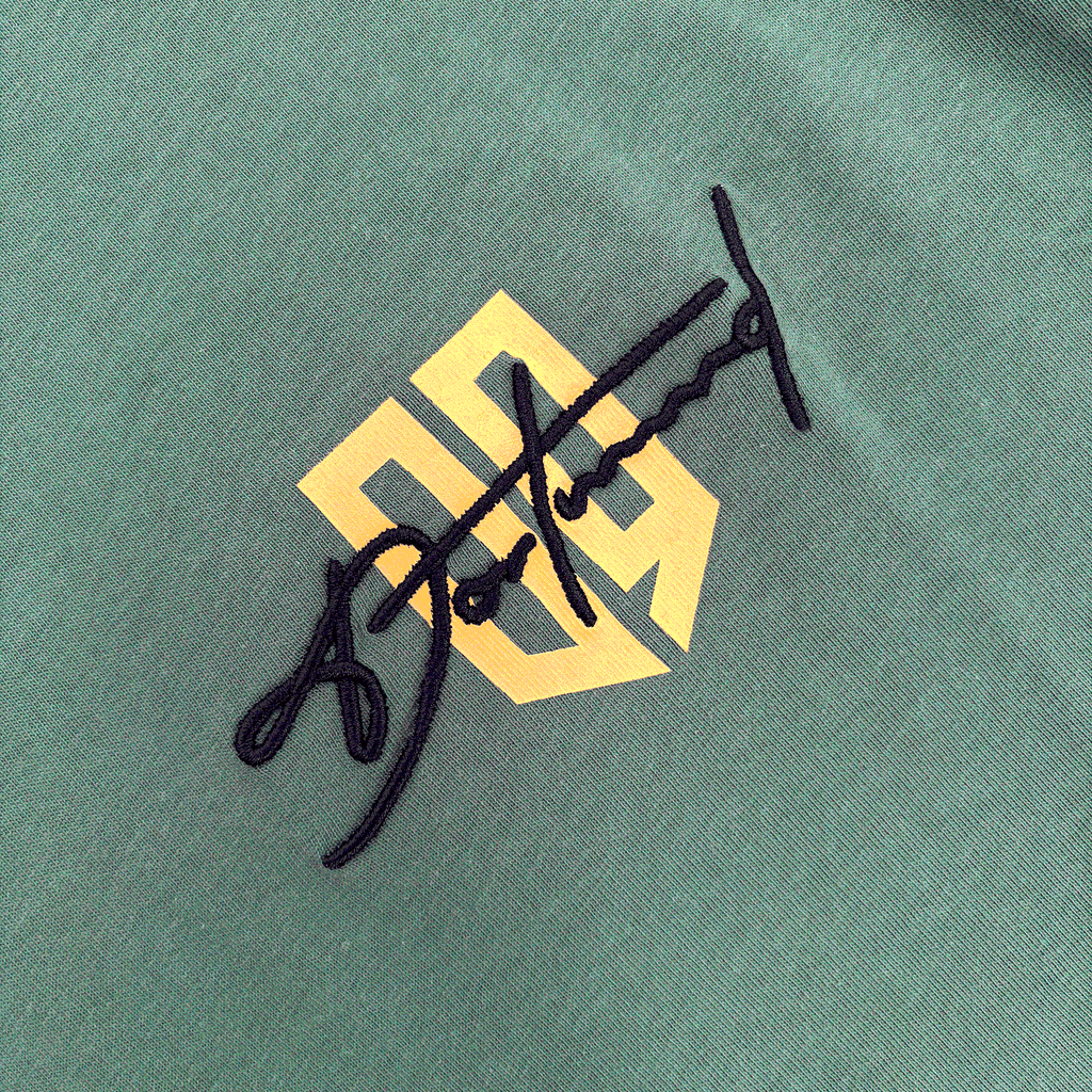 Nahaufnahme eines BVB T-Shirts Lifestyle in Duckgreen mit prominentem gelben BVB-Logo und schwarzer Signaturstickerei.