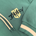 Dieser duckgrüne BVB-Hoodie zeigt das Borussia Dortmund-Logo in schwarzgelb, beige Kordeln und einen gestreiften Ärmelabschluss.