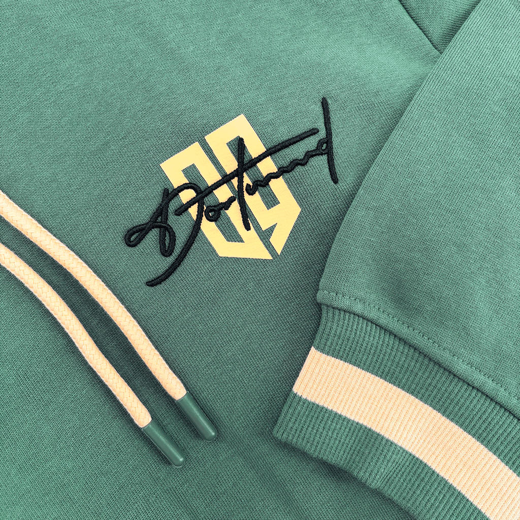 Dieser duckgrüne BVB-Hoodie zeigt das Borussia Dortmund-Logo in schwarzgelb, beige Kordeln und einen gestreiften Ärmelabschluss.