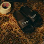 Black BVB bath slippers with white lettering that reads “Art of Defense | Inspired by Nico,” displayed on a colorful, patterned rug.