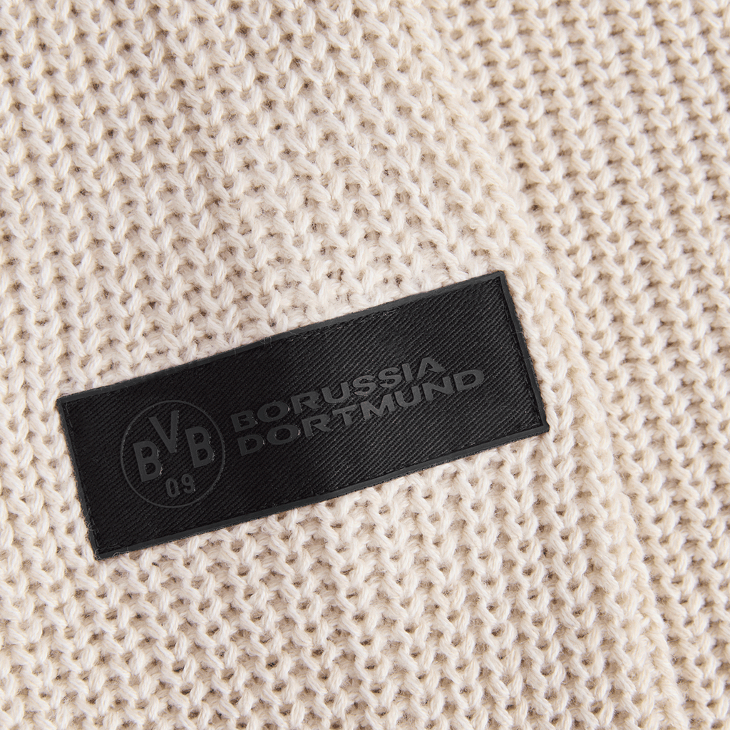 Beiger BVB-Winterpullover in Nahaufnahme, mit schwarzem Borussia Dortmund-Logo auf einem Etikett auf der Vorderseite des Strickstoffs.