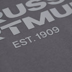 Detailaufnahme eines grauen BVB Urban T-Shirts mit großem weißen Schriftzug 'EST. 1909' und sichtbaren Elementen des Vereinsnamens Borussia Dortmund.