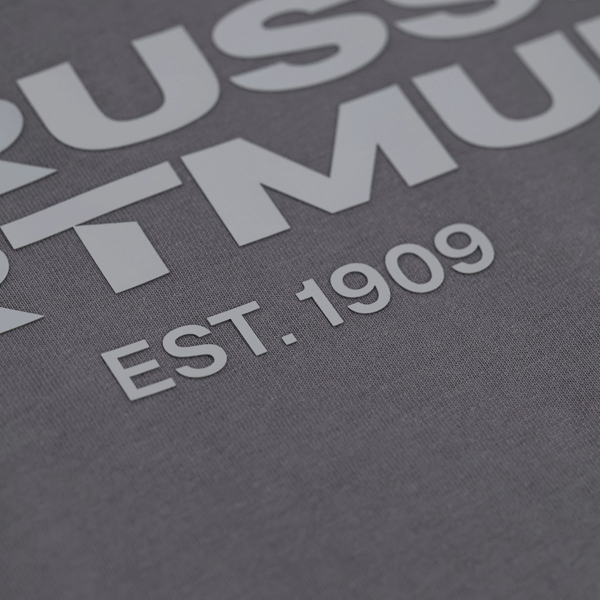 Detailaufnahme eines grauen BVB Urban T-Shirts mit großem weißen Schriftzug 'EST. 1909' und sichtbaren Elementen des Vereinsnamens Borussia Dortmund.
