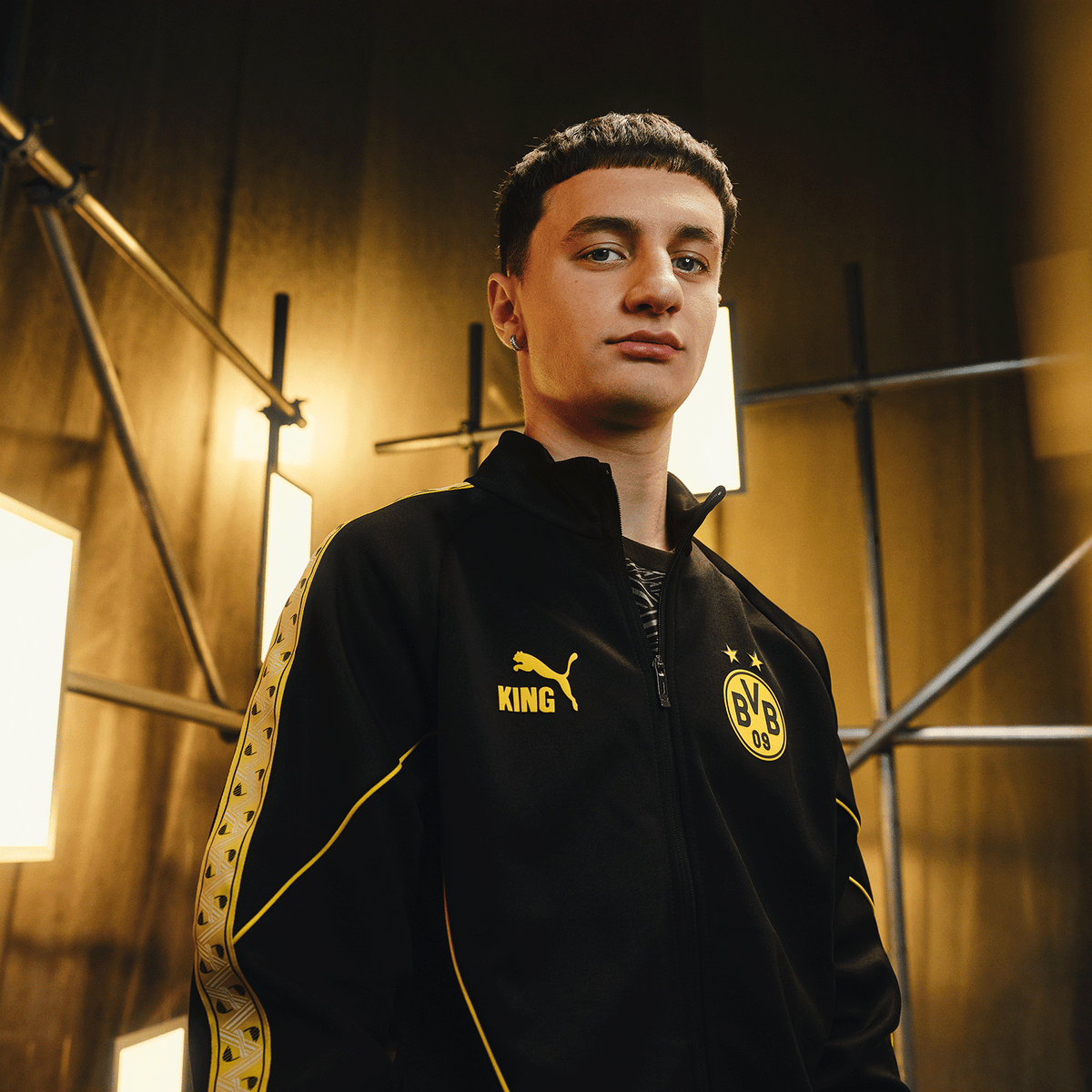Black-and-yellow BVB King Anthem jacket from the 26-27 collection, featuring the BVB logo, the PUMA logo, and "KING" lettering, presented by a person in front of a yellow-lit background.