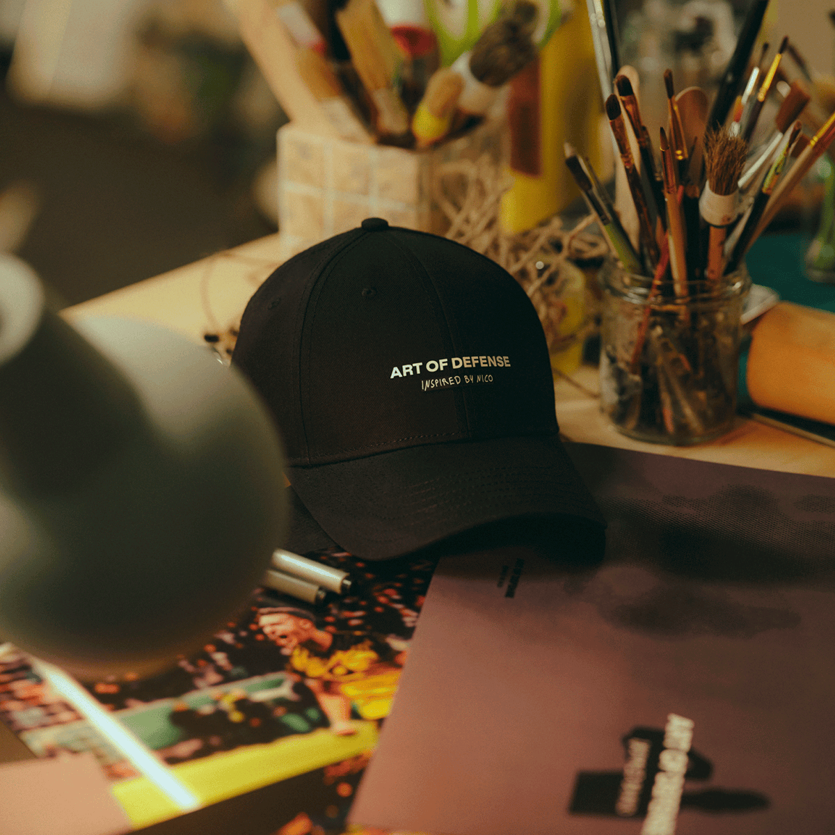 Cappellino nero del BVB con la scritta bianca "Art of Defense - Inspired by Nico", appoggiato su un tavolo e circondato da materiale per dipingere.