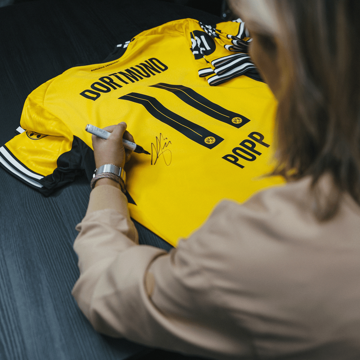 Ein gelbes BVB-Trikot mit der Aufschrift 'Dortmund' und 'Popp' wird auf einem schwarzen Tisch von einer Person signiert. Sichtbar sind Details des Trikots und die Rückansicht.