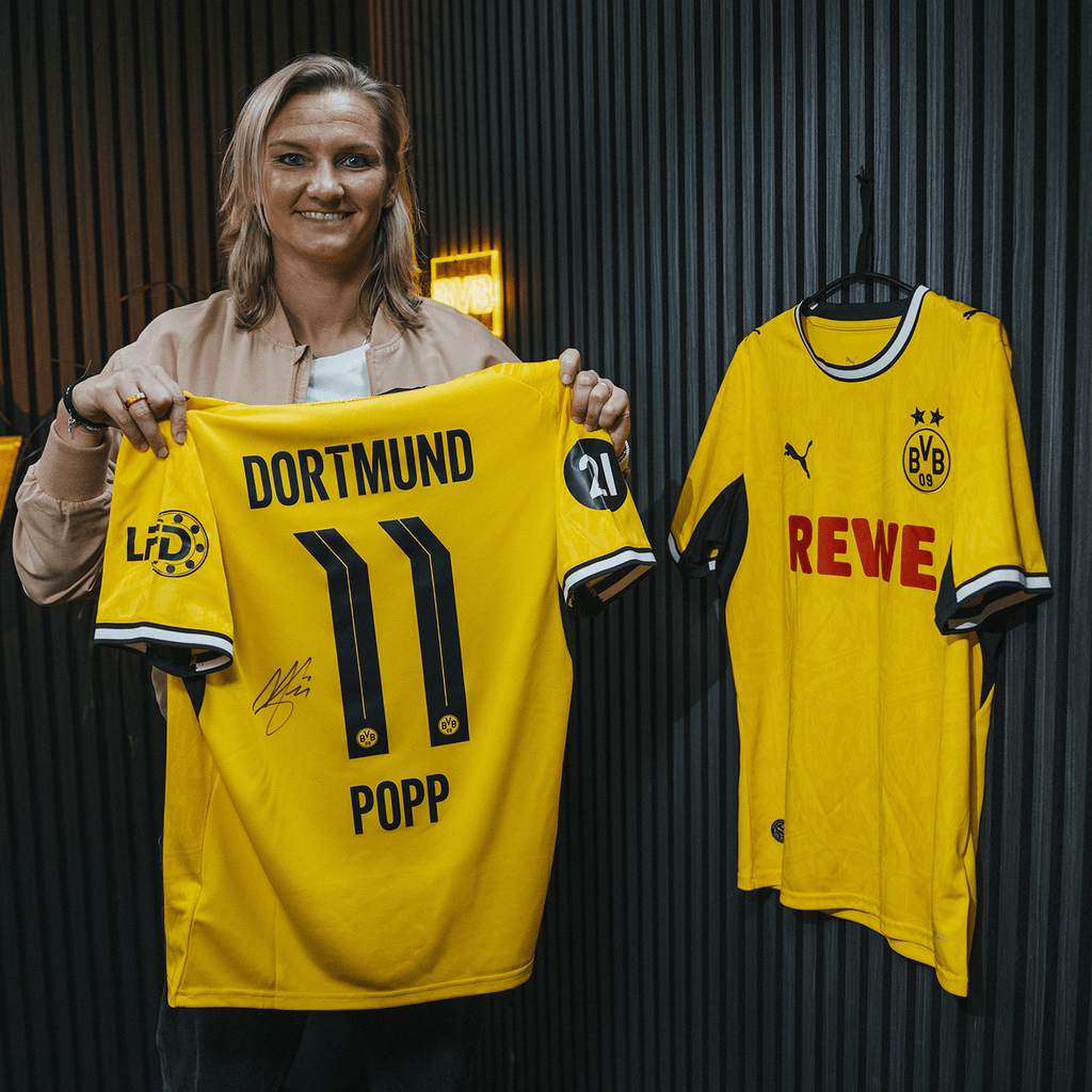 Eine Person präsentiert ein gelbes BVB-Trikot mit der Rückseite der Nummer 11 und dem Namen Popp. Neben ihr hängt ein weiteres BVB-Trikot mit REWE-Logo an einer Wand.