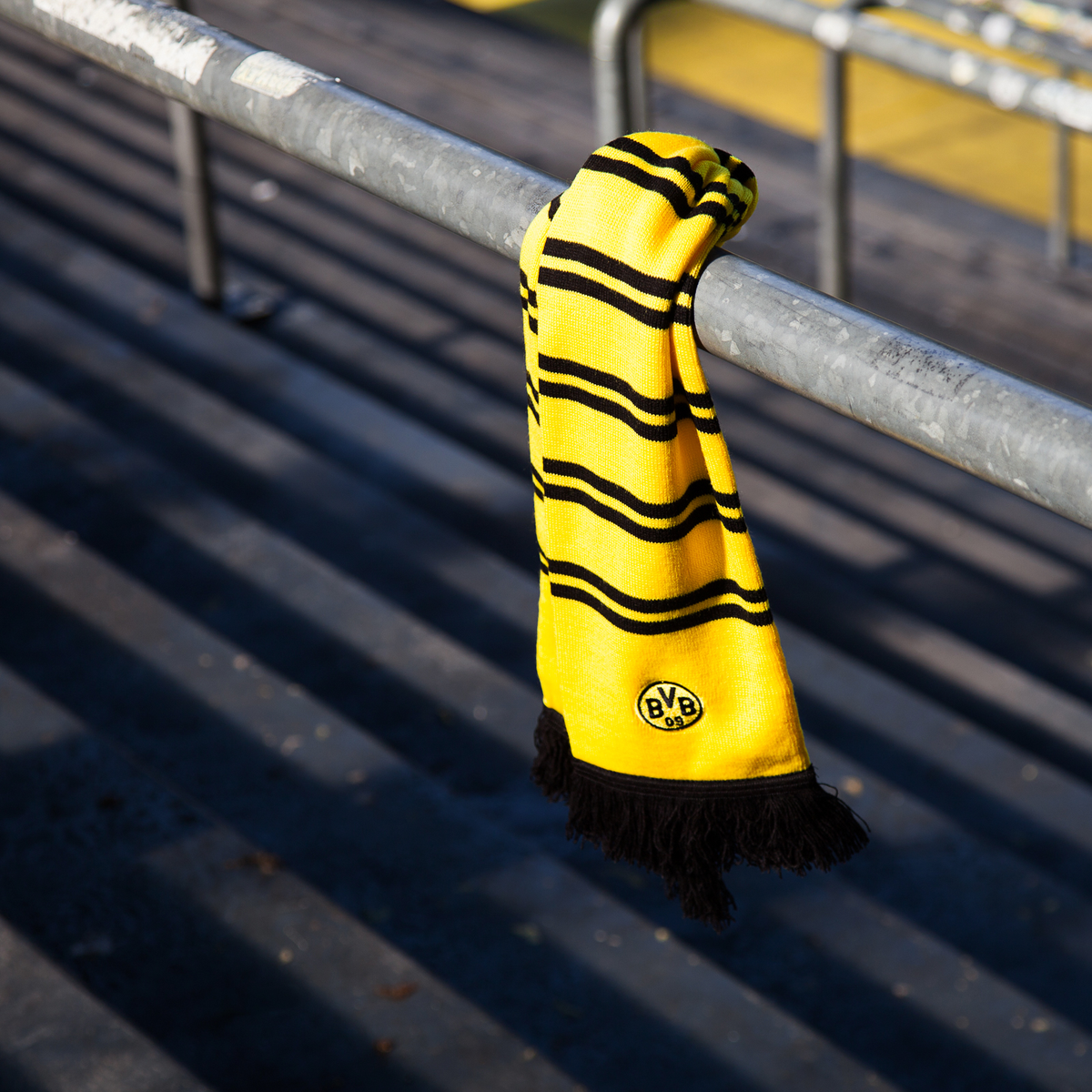 Stylischer BVB-Schal in schwarzgelbem Design mit Ringelstreifen, schwarzem Fransenabschluss und prominentem BVB-Emblem, fotografiert im Stadion auf einem Geländer.